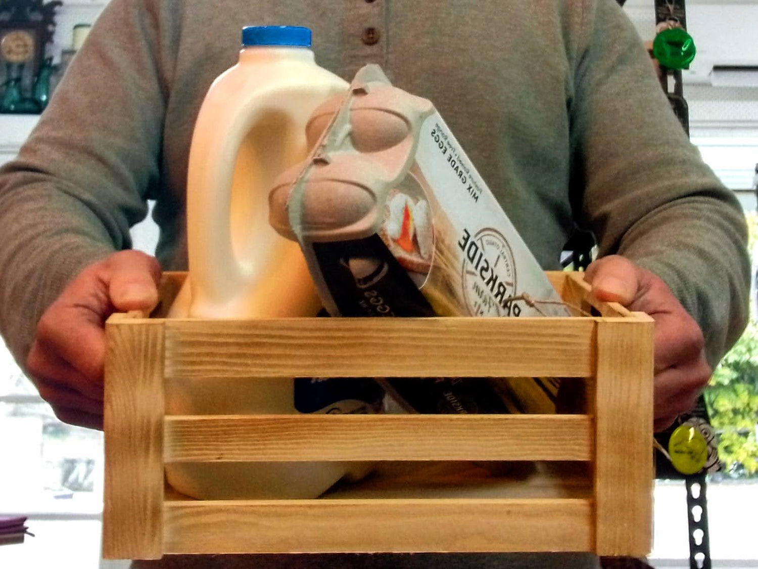 Person holding a wooden crate with a bottle of milk, and a carton of eggs.